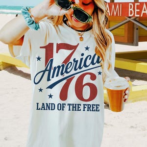May include: Cream-coloured t-shirt with the text "17 America 76 Land of the Free" in red and blue. The shirt is worn with a pink cap and sunglasses. A beverage in a clear cup is held in the hand.