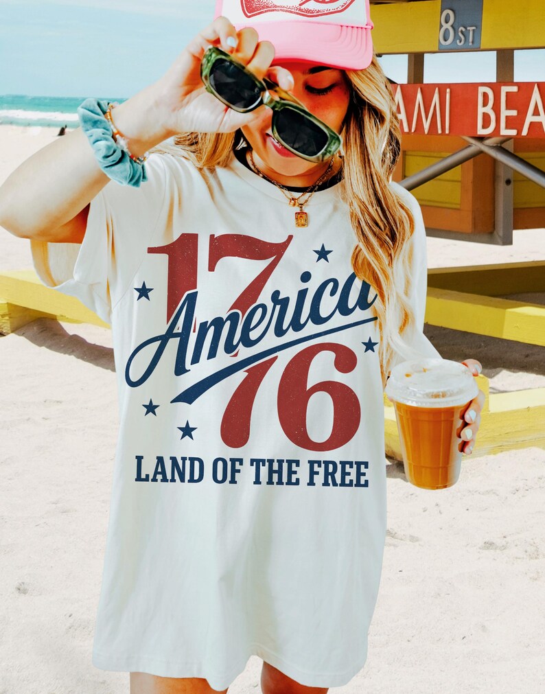 May include: Cream-coloured t-shirt with the text "17 America 76 Land of the Free" in red and blue. The shirt is worn with a pink cap and sunglasses. A beverage in a clear cup is held in the hand.