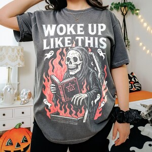 May include: Dark grey t-shirt featuring the text "WOKE UP LIKE THIS" and a graphic of a skeleton in a hooded robe reading a book amidst flames. The design includes ghosts and a coffin. The shirt has a relaxed, oversized fit.