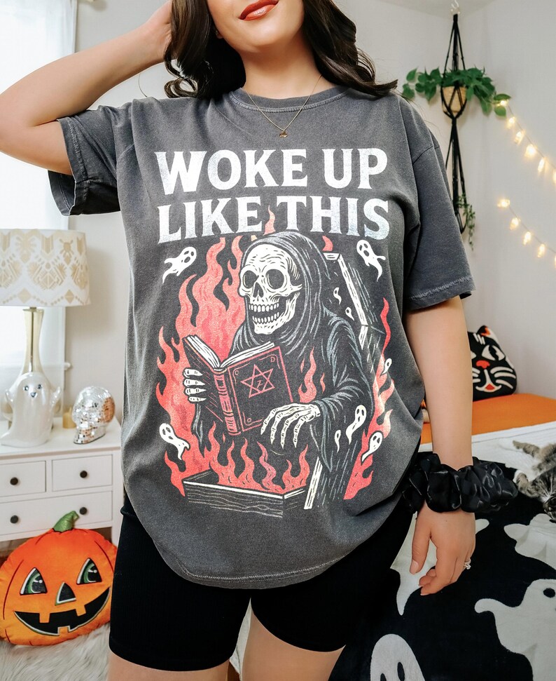 May include: Dark grey t-shirt featuring the text "WOKE UP LIKE THIS" and a graphic of a skeleton in a hooded robe reading a book amidst flames. The design includes ghosts and a coffin. The shirt has a relaxed, oversized fit.