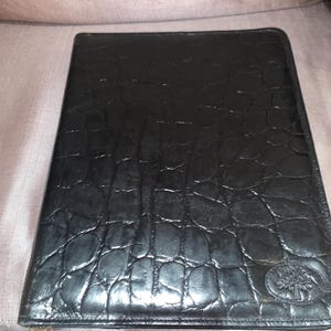 May include: Black leather portfolio with a textured crocodile pattern. The portfolio has a zipper closure and a small embossed logo of a tree. The leather appears to be high-quality and the portfolio is rectangular in shape.