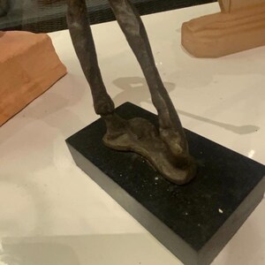 Puede incluir: Una escultura de bronce de patas de animal, montada sobre una base rectangular negra. Las patas tienen una superficie texturizada, que sugiere m&uacute;sculo y forma. La escultura est&aacute; sobre una superficie blanca.