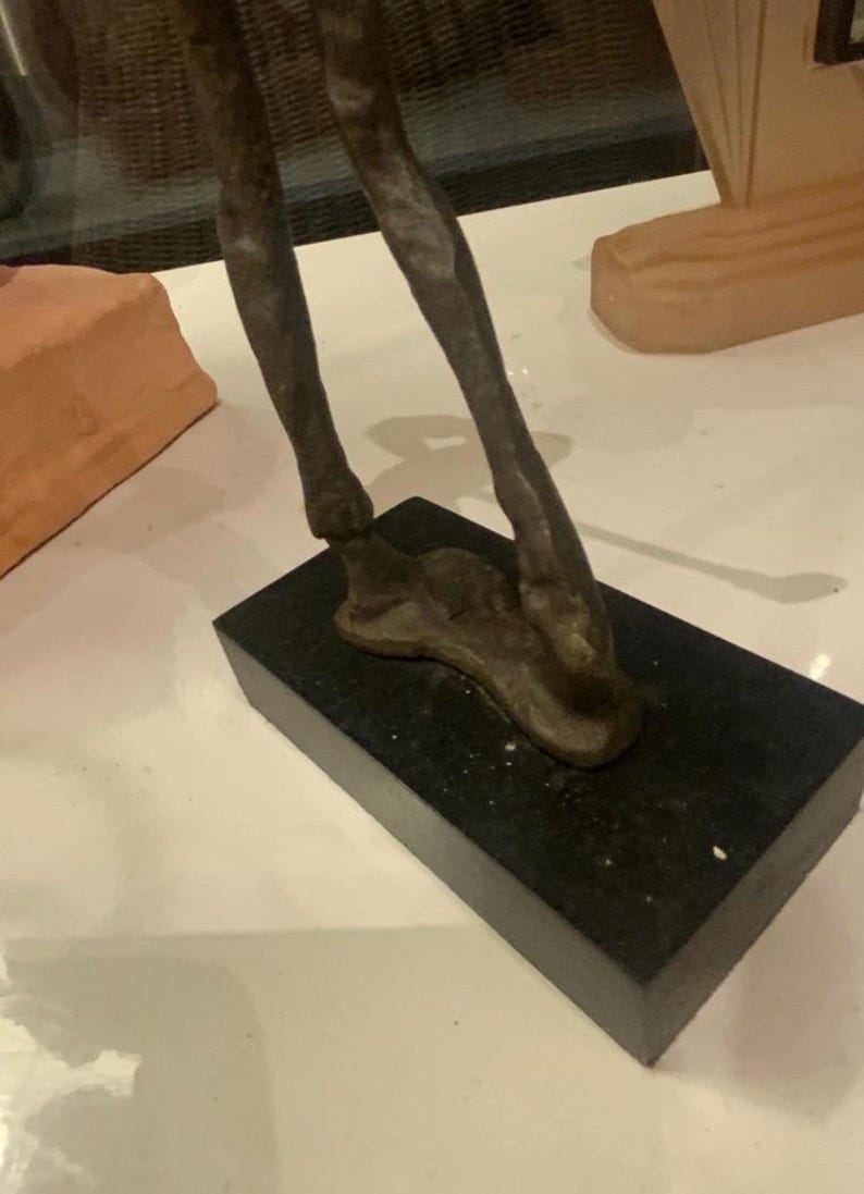 Puede incluir: Una escultura de bronce de patas de animal, montada sobre una base rectangular negra. Las patas tienen una superficie texturizada, que sugiere m&uacute;sculo y forma. La escultura est&aacute; sobre una superficie blanca.