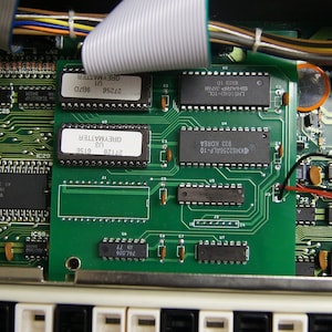 May include: A detailed view of a green circuit board populated with black integrated circuits and other electronic components. Some chips are labelled with "GREYMATTER" and other codes. A white ribbon cable and keyboard keys are also visible.