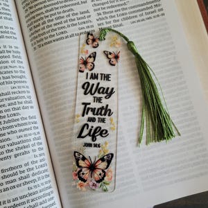 May include: Clear acrylic bookmark featuring the text "I AM THE WAY THE TRUTH AND THE LIFE" with John 14:6. The bookmark has butterfly and floral designs, a green tassel, and is placed on an open book.