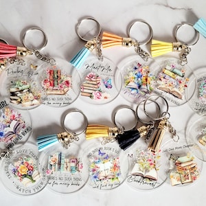 May include: A collection of clear acrylic keychains with silver key rings and colorful tassels. Each keychain features a book-themed design with floral accents and phrases like "Just a Girl Who Loves Books" and "Meet Me".