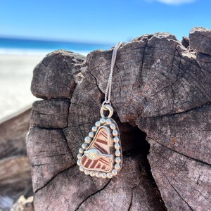 Pode incluir: Um colar com pingente de prata com um pingente triangular único. O pingente apresenta um design marrom e branco, emoldurado por uma borda prateada com contas. Um pequeno detalhe de folha é visível no pingente. O colar é exibido em uma superfície de madeira envelhecida.