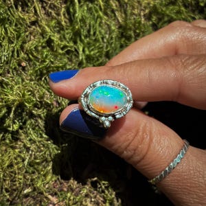 May include: Oval opal ring with a silver filigree setting. The opal displays a vibrant array of colors, including blue, green, orange, and red. The ring is held up against a background of green moss.