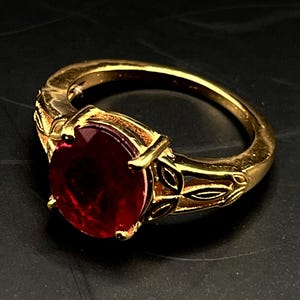 Könnte beinhalten: Ein goldfarbener Ring mit einem großen, oval geschliffenen, tiefroten Edelstein. Der Ring hat ein verziertes Design mit blattartigen Details am Band und um die Fassung des Edelsteins. Der Ring ist vor einem dunklen Hintergrund platziert.