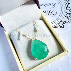 Puede incluir: Un collar de oro con un gran colgante de piedra verde facetada en forma de pera. El collar se exhibe en una caja crema abierta, con interior burdeos. La joya es delicada y elegante, ideal para ocasiones especiales.