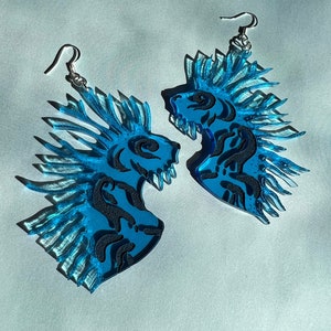 May include: A pair of blue acrylic earrings featuring a stylized lion design with black detailing. The earrings have a textured, layered appearance and are suspended from silver-colored fish hook ear wires. The earrings are displayed against a light blue background.