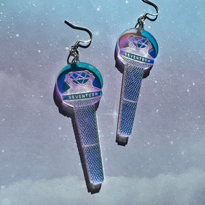 May include: A pair of iridescent silver earrings featuring a microphone design with the word "SEVENTEEN" printed on the top. The microphone is a clear plastic with a holographic finish.