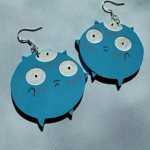 May include: A pair of blue, round, wooden earrings with a cartoon face and three eyes. The earrings have silver hooks.
