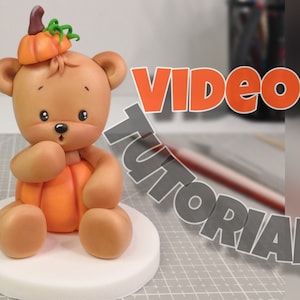 May include: A brown sugar paste teddy bear wearing a pumpkin hat and holding a pumpkin. The bear is sitting on a white round base. The text "Sugar paste decor VIDEO TUTORIAL" is in the background.