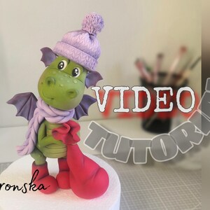 May include: A green dragon wearing a purple hat and scarf, holding a red bag. The dragon is made of sugar paste and is a cake topper. The text "SUGAR PASTE DECOR by Vronska VIDEO TUTORIAL" is visible in the background.