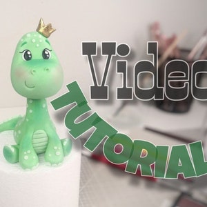 May include: A green sugar paste dinosaur figurine with a gold crown, sitting on a white cake base. The text "Sugar paste decor Video Tutorial" is visible in the background.