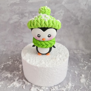 May include: A green and white penguin cake topper wearing a green knit hat and scarf. The penguin is standing on a white cake base.
