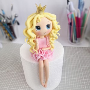 May include: A fondant cake topper of a princess doll with blonde curly hair, wearing a pink dress and a gold crown. The doll is sitting on a white cake stand.