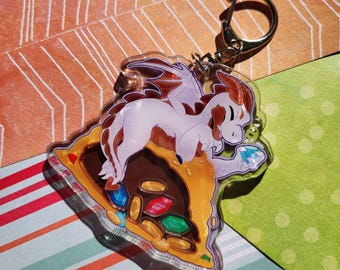 May include: A clear acrylic keychain featuring a cartoon dragon holding a blue gem. The dragon is sitting on a pile of gold coins and colorful gems. The keychain has a gold metal ring.