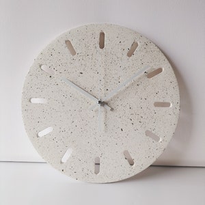 Op de afbeelding: Ronde wandklok in gebroken wit met een gespikkelde, terrazzo-achtige wijzerplaat. De klok heeft zilveren wijzers en langwerpige uitsparingen als uurmarkeringen. Diameter ongeveer 30 cm. Modern, minimalistisch ontwerp.