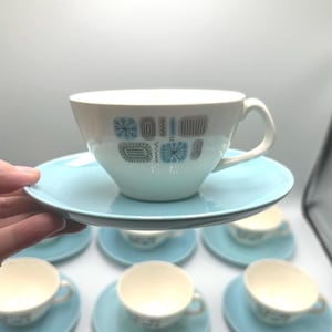 May include: A white ceramic teacup and saucer set featuring a mid-century modern pattern of blue and grey geometric shapes and starbursts. The teacup has a curved handle, and the saucer is a solid light blue color with a matching white center.