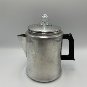 May include: A vintage aluminum stovetop percolator coffee pot featuring a black heat-resistant handle, a triangular spout, and a circular lid topped with a clear glass knob. The metallic body shows signs of wear and has visible measurement markings.