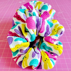 May include: A colourful hair scrunchie with a white background and a pattern of yellow, purple, pink and blue peeps. The scrunchie is made of a soft, stretchy fabric.