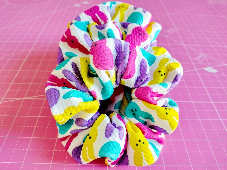 May include: A colourful hair scrunchie with a white background and a pattern of yellow, purple, pink and blue peeps. The scrunchie is made of a soft, stretchy fabric.