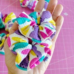 May include: A pair of colourful hair scrunchies with a white background and a pattern of purple, pink, yellow, and turquoise shapes. The scrunchies are made from a soft, stretchy fabric.