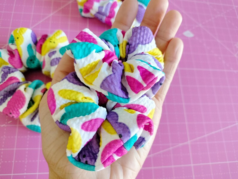 May include: A pair of colourful hair scrunchies with a white background and a pattern of purple, pink, yellow, and turquoise shapes. The scrunchies are made from a soft, stretchy fabric.