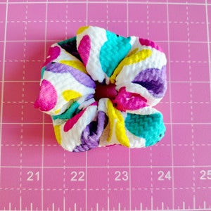 May include: A colourful hair scrunchie with a white background and a pattern of pink, purple, yellow, and turquoise circles. The scrunchie is made of a soft, textured fabric.