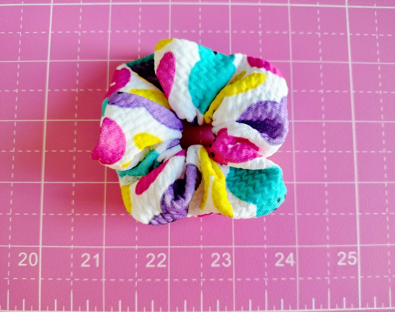 May include: A colourful hair scrunchie with a white background and a pattern of pink, purple, yellow, and turquoise circles. The scrunchie is made of a soft, textured fabric.