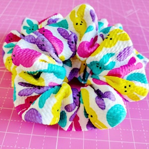 May include: A white scrunchie with a colourful pattern of yellow, pink, purple, and turquoise bunnies. The bunnies have black eyes and a simple smile.
