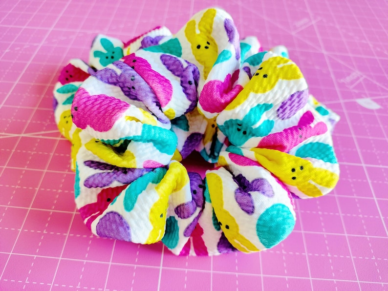 May include: A white scrunchie with a colourful pattern of yellow, pink, purple, and turquoise bunnies. The bunnies have black eyes and a simple smile.