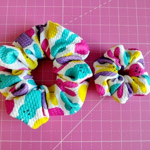 May include: Two colourful hair scrunchies with a white background and a pattern of pastel pink, yellow, and turquoise shapes. The scrunchies are made of a soft, textured fabric.