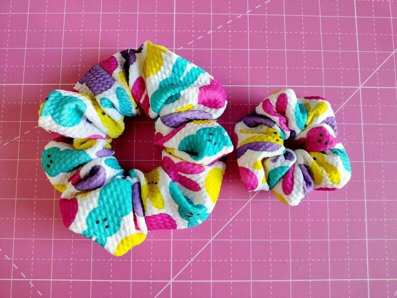 May include: Two colourful hair scrunchies with a white background and a pattern of pastel pink, yellow, and turquoise shapes. The scrunchies are made of a soft, textured fabric.