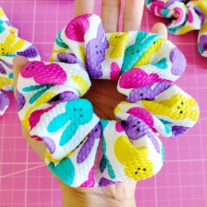 May include: A white scrunchie with a colourful pattern of pastel yellow, pink, purple, and blue bunnies. The bunnies have black eyes and a single black button nose.