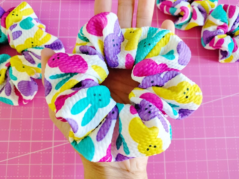 May include: A white scrunchie with a colourful pattern of pastel yellow, pink, purple, and blue bunnies. The bunnies have black eyes and a single black button nose.
