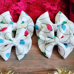 May include: Four white fabric hair bows with a Christmas light pattern. The lights are red, green, blue, and yellow.