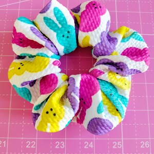 May include: A white scrunchie with a colourful pattern of yellow, pink, purple and blue Peeps. The Peeps are in the shape of bunnies and chicks.