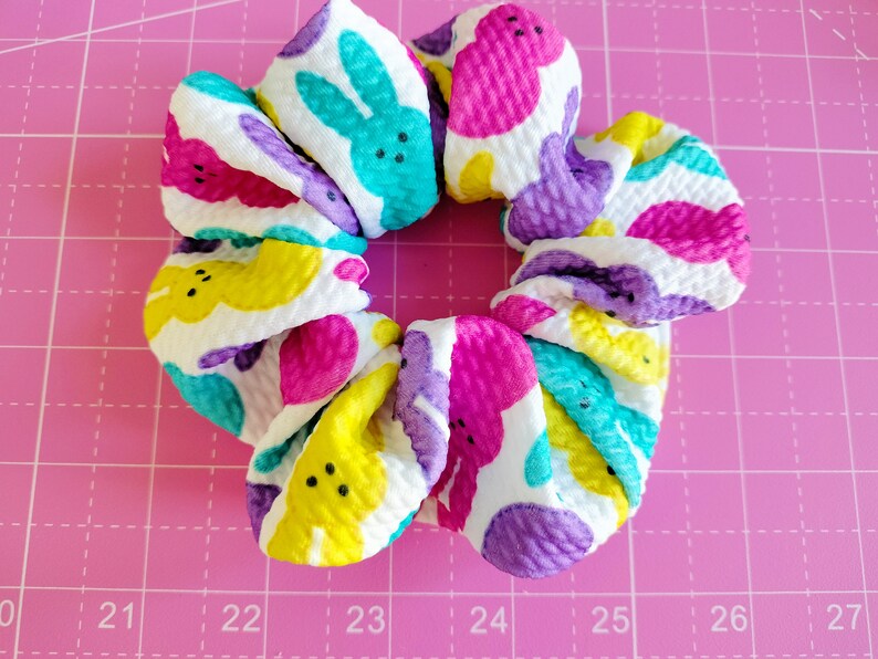 May include: A white scrunchie with a colourful pattern of yellow, pink, purple and blue Peeps. The Peeps are in the shape of bunnies and chicks.
