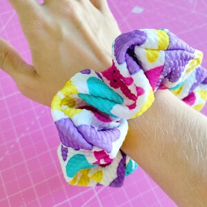 May include: A colourful scrunchie with a white background and purple, yellow, pink, and turquoise accents. The scrunchie is made of a soft, textured fabric.