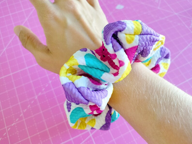 May include: A colourful scrunchie with a white background and purple, yellow, pink, and turquoise accents. The scrunchie is made of a soft, textured fabric.