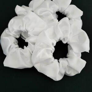 May include: Four white satin scrunchies, each with a gathered, ruffled design. The scrunchies are arranged in a pile on a dark green background.