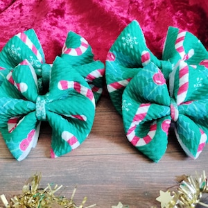 May include: Two green and white Christmas-themed hair bows with candy cane and holly designs.