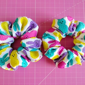 May include: Two colourful hair scrunchies with a white background and a pattern of pastel-coloured Peeps. The scrunchies are made of a soft, textured fabric.