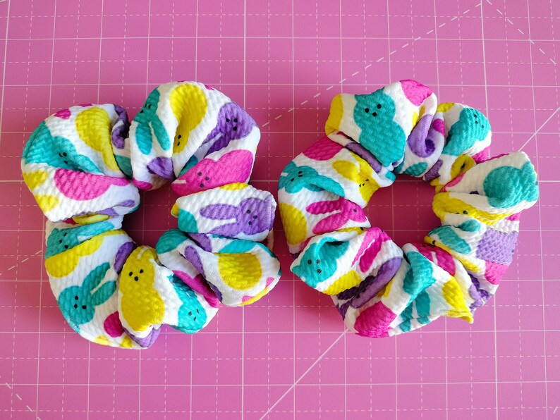 May include: Two colourful hair scrunchies with a white background and a pattern of pastel-coloured Peeps. The scrunchies are made of a soft, textured fabric.