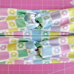May include: A pastel checkered headband with a knotted design. The headband is made of a soft, stretchy fabric and features a repeating pattern of pastel pink, yellow, green, and blue squares with white flowers.