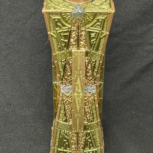 Könnte beinhalten: Eine goldene und grüne Metall-Rätselbox mit komplizierten Designs. Die Box ist geschlossen und hat eine kreisförmige Öffnung oben.