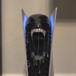 May include: A white gaming console with a black alien head on the front. The alien head is dripping with a white substance, and the console is lit up with blue lights.